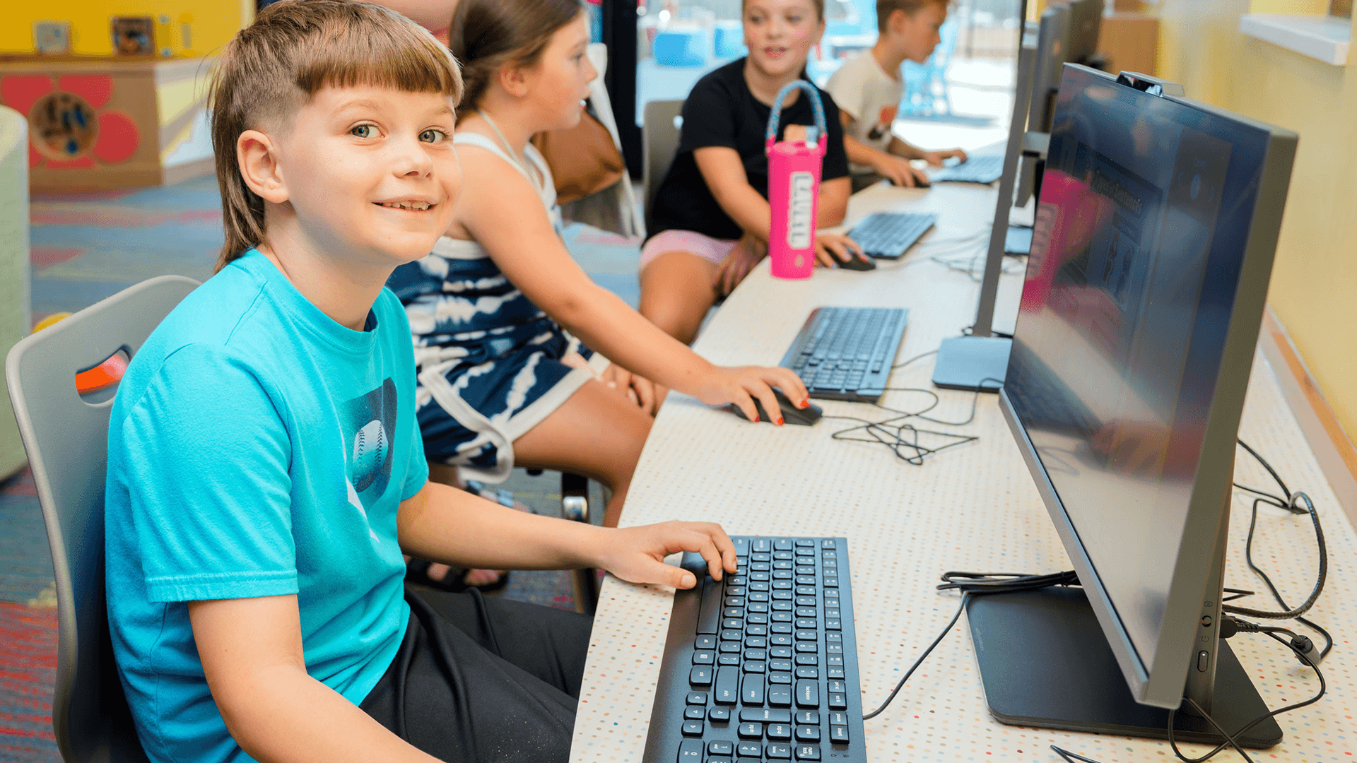 Children using desktop computers in a computer lab, with one child smiling at the camera.