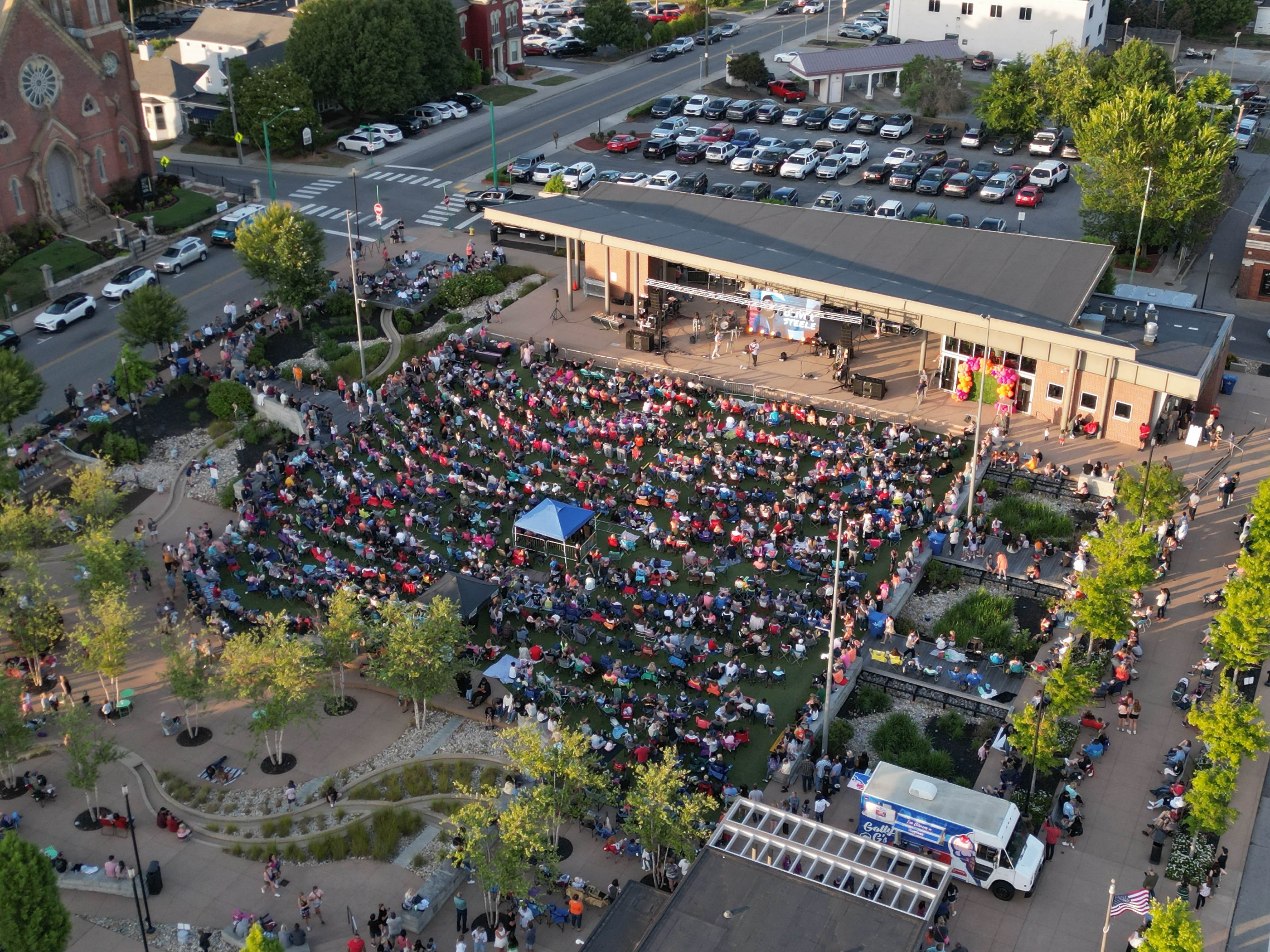 A large crowd gathered around a concert stage in a downtown park.